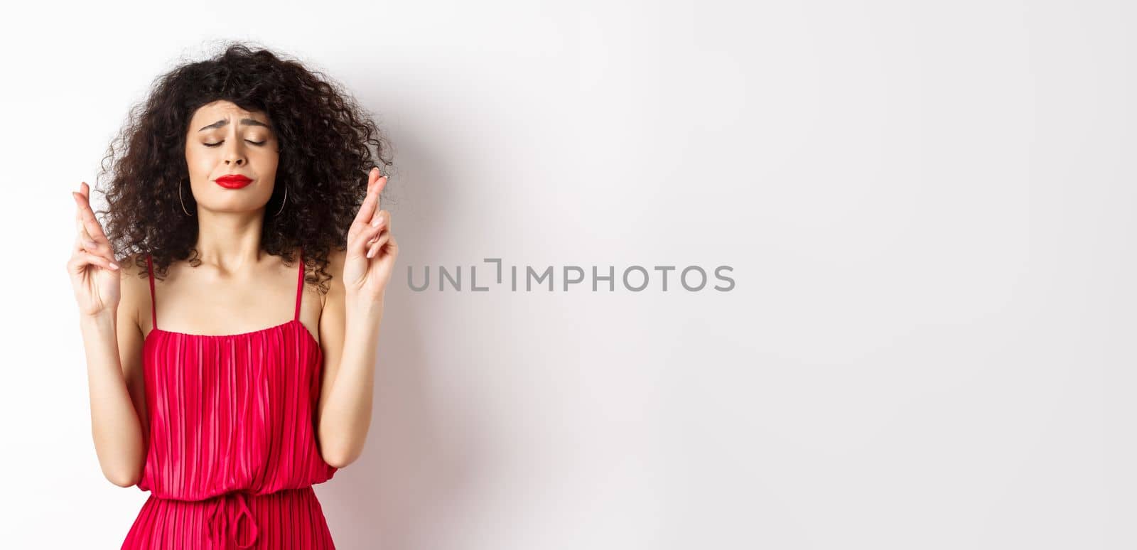 Hopeful woman in red dress making wish, praying with fingers crossed and closed eyes, pleading god, standing over white background by Benzoix