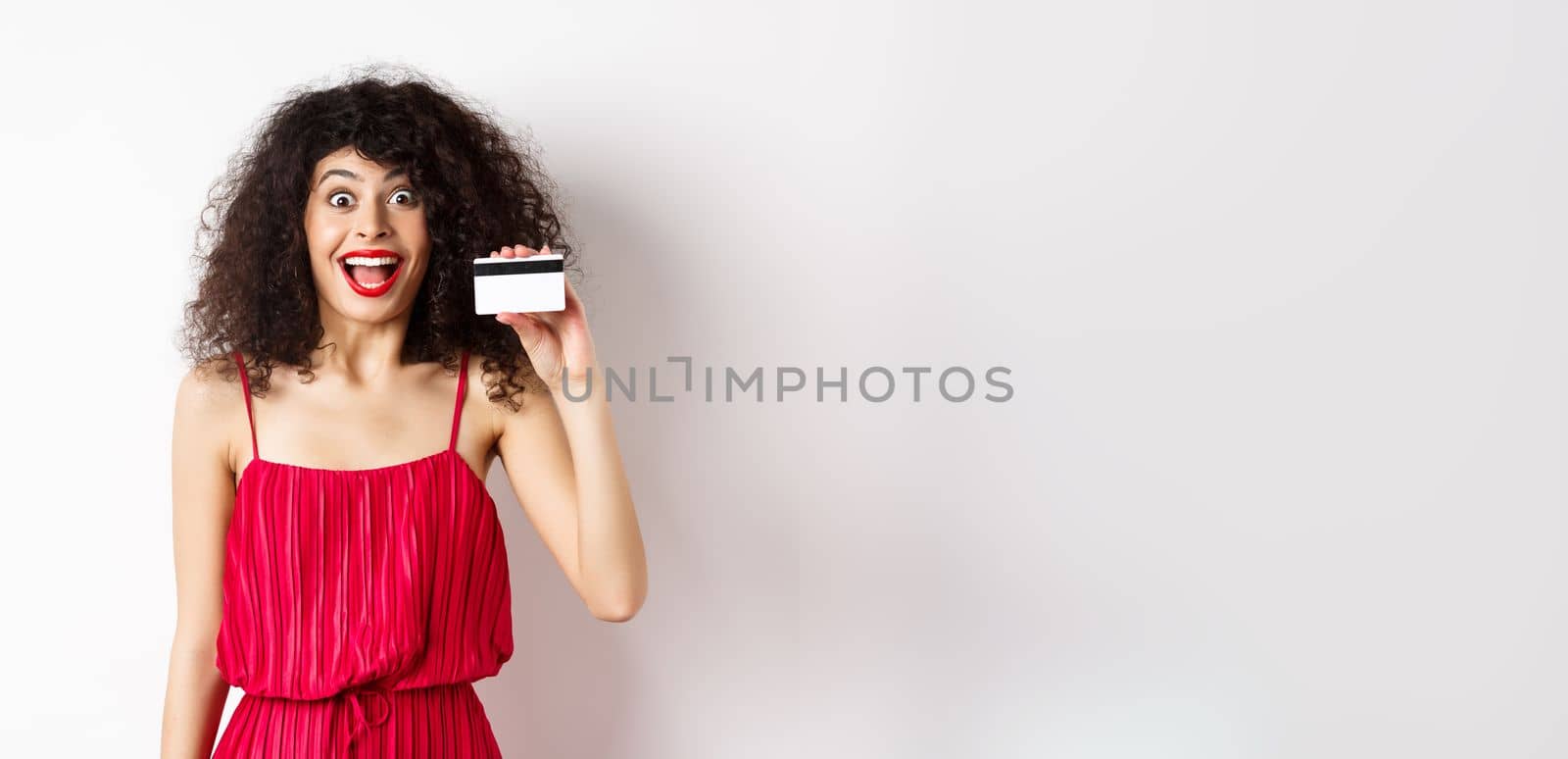 Excited smiling woman in red dress, showing plastic credit card and looking happy, standing over white background. Copy space