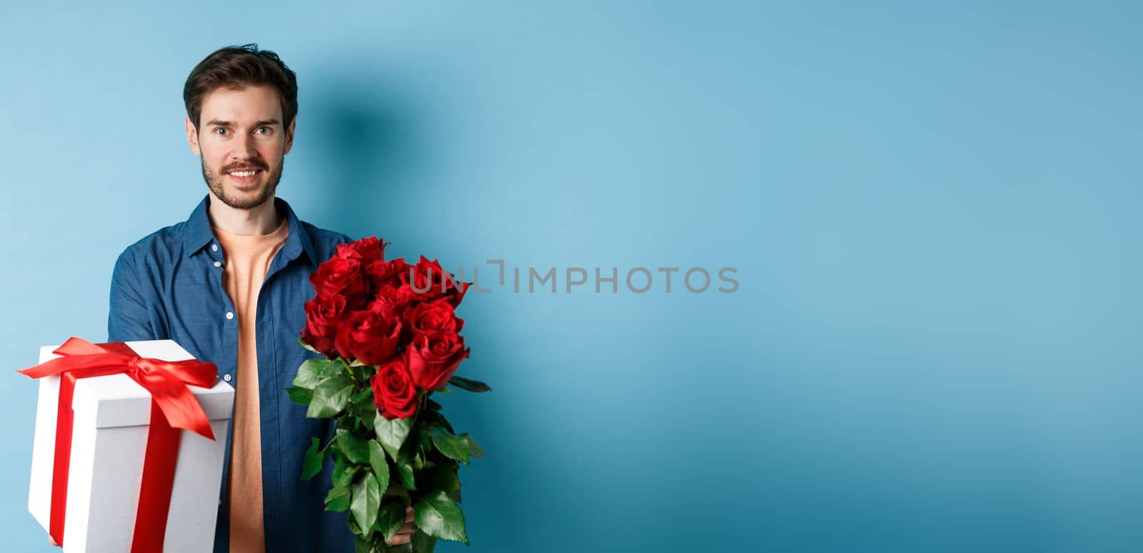 Love and Valentines day concept. Charming young man giving gift and bouquet of roses to girlfriend, standing over blue background.