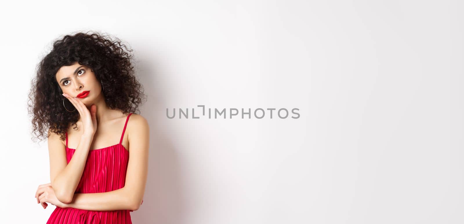 Annoyed and bored young woman with curly hair, look away distressed, lean face on hand, standing bothered in red dress on white background.