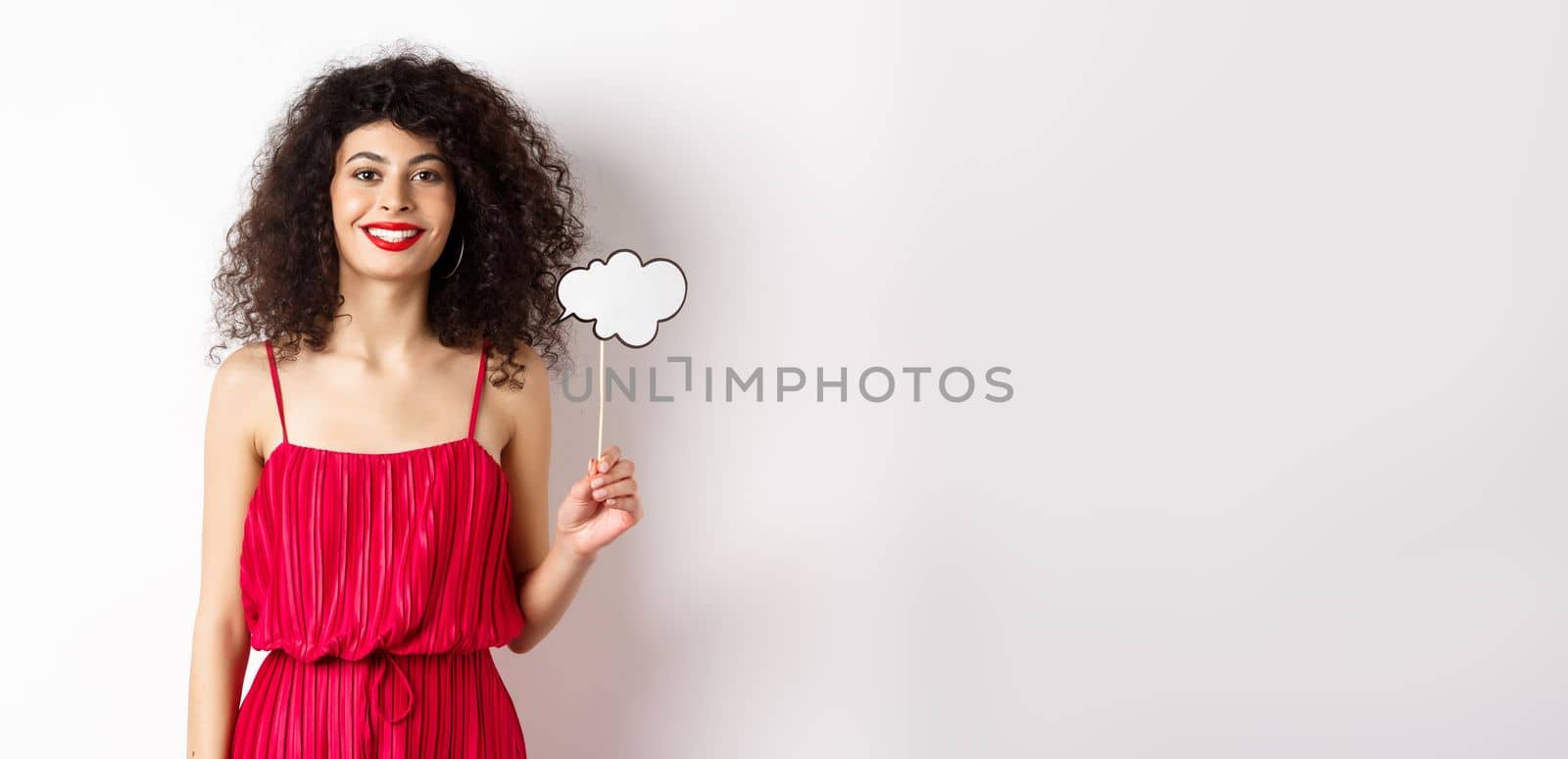 Happy stylish woman with curly hair, beauty makeup, holding comment cloud on stick and smiling, standing in red dress on white background by Benzoix