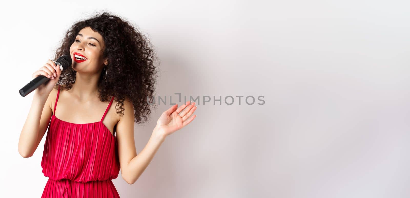 Elegant curly-haired woman in red dress singing in microphone, looking aside and smiling happy, standing on white background.