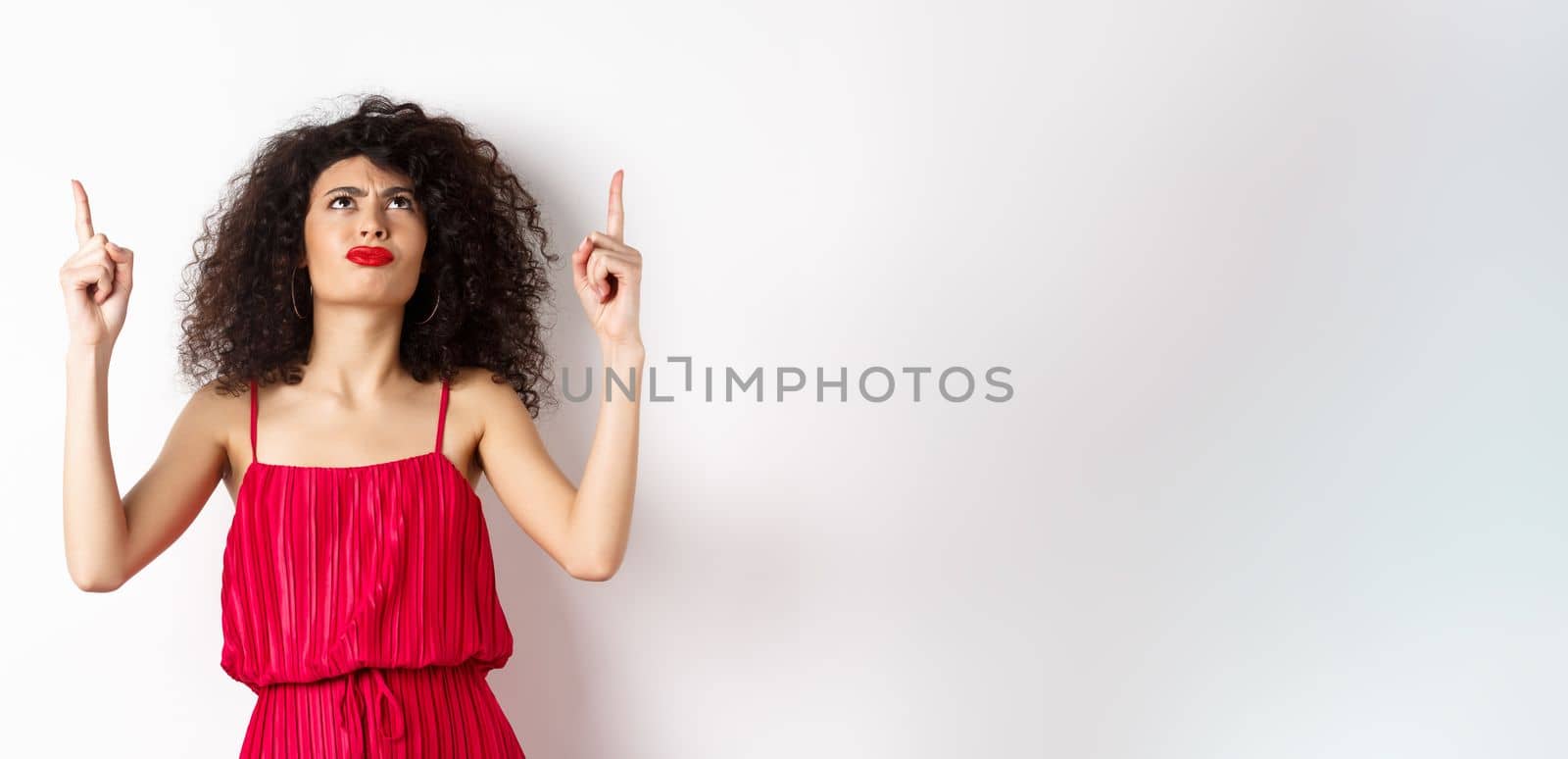 Angry and grumpy woman with curly hair, wearing red dress, frowning and looking up disappointed, standing over white background.