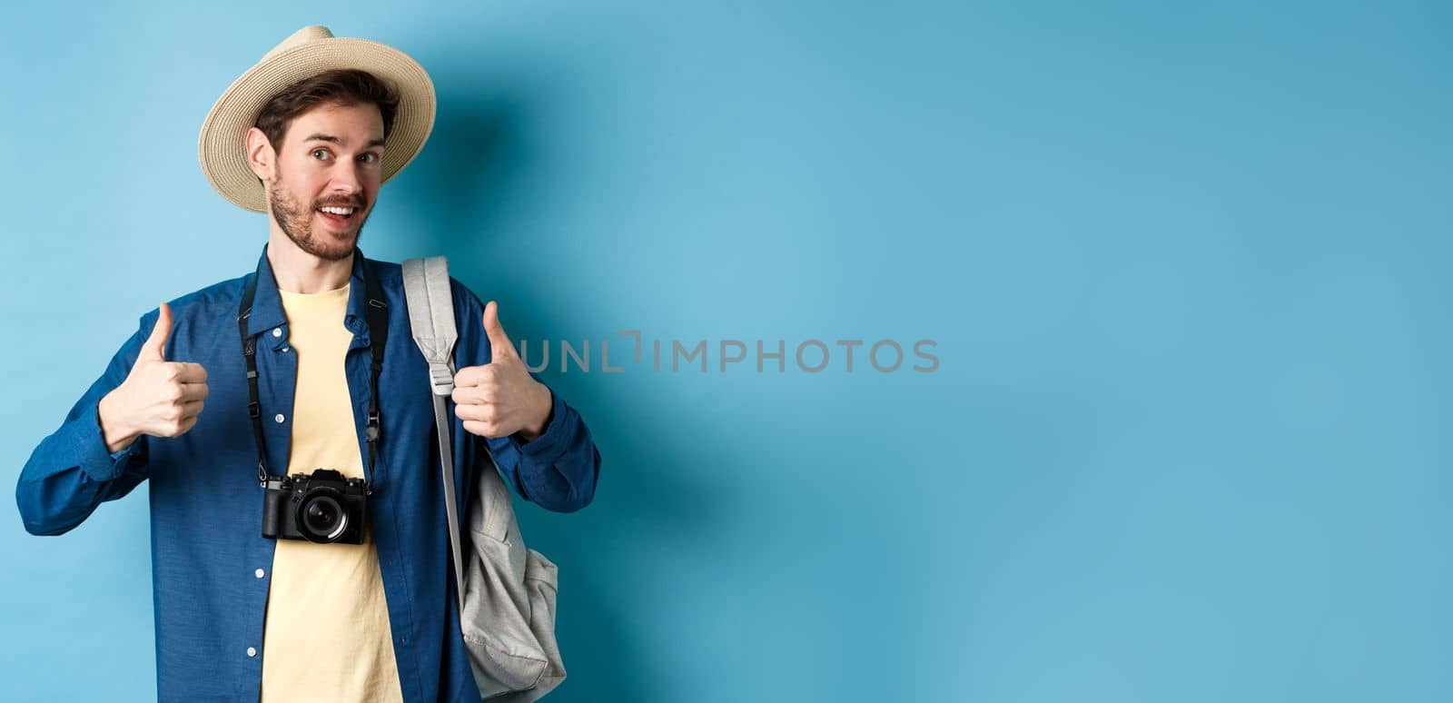 Happy smiling tourist say yes and show thumbs up, going on summer vacation, holding backpack and photo camera, blue background.