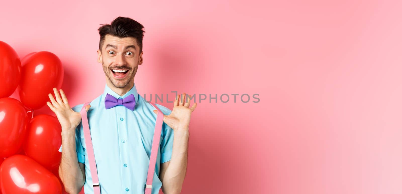 Valentines day concept. Happy young man looking surprised, raising hands up joyful, celebrating near big red hearts and pink background by Benzoix