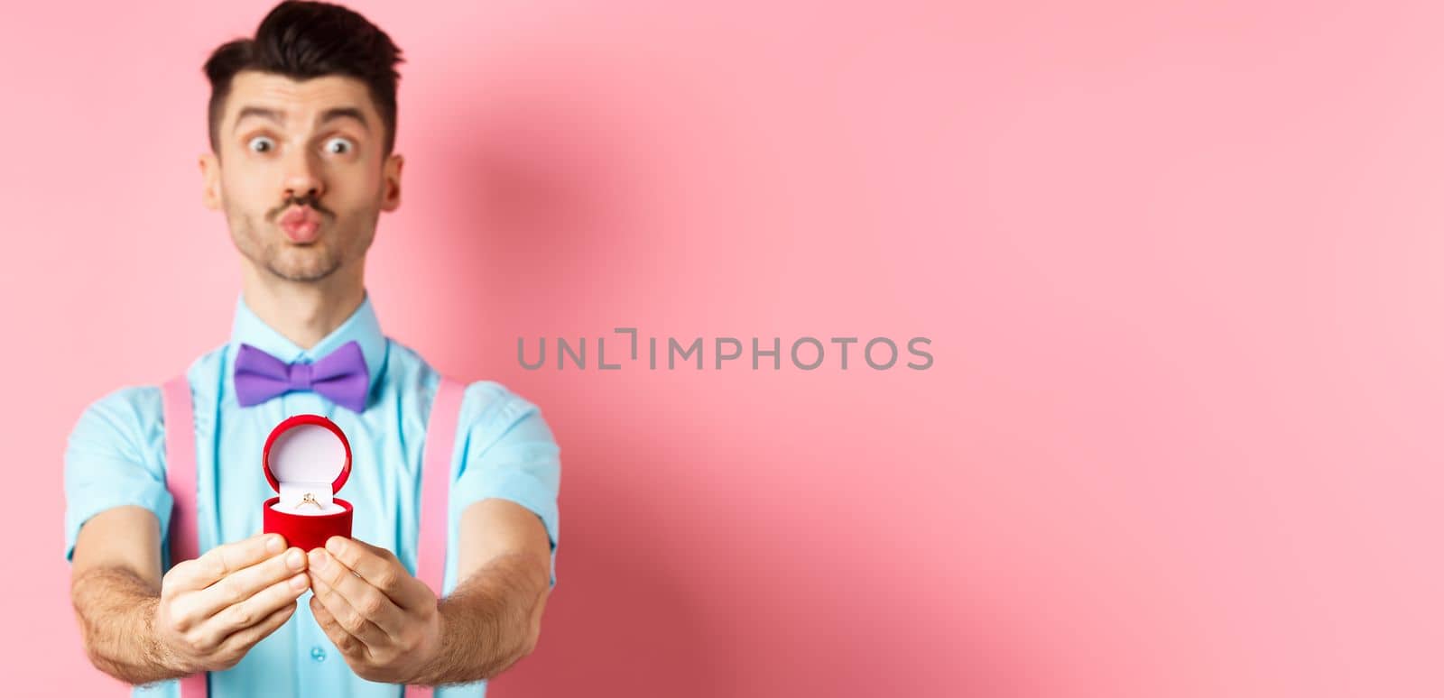 Valentines day. Funny young man pucker lips for kiss and showing engagement ring, making proposal, say marry me to lover, standing over pink background by Benzoix