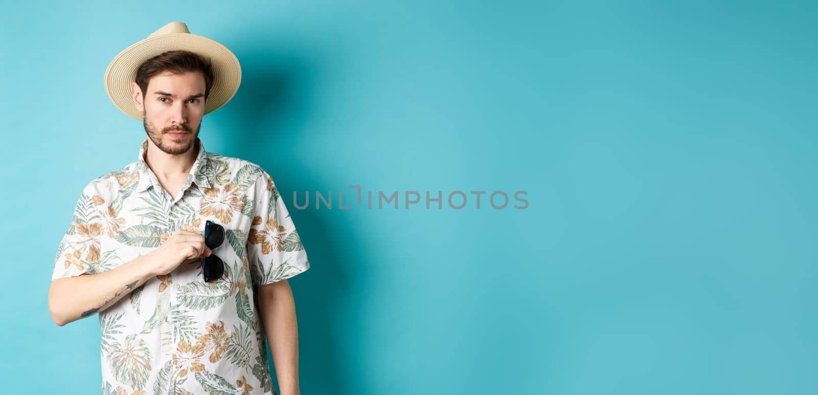 Handsome tourist in hawaiian shirt and summer hat, put sunglasses in pocket, going on vacation, standing against blue background.