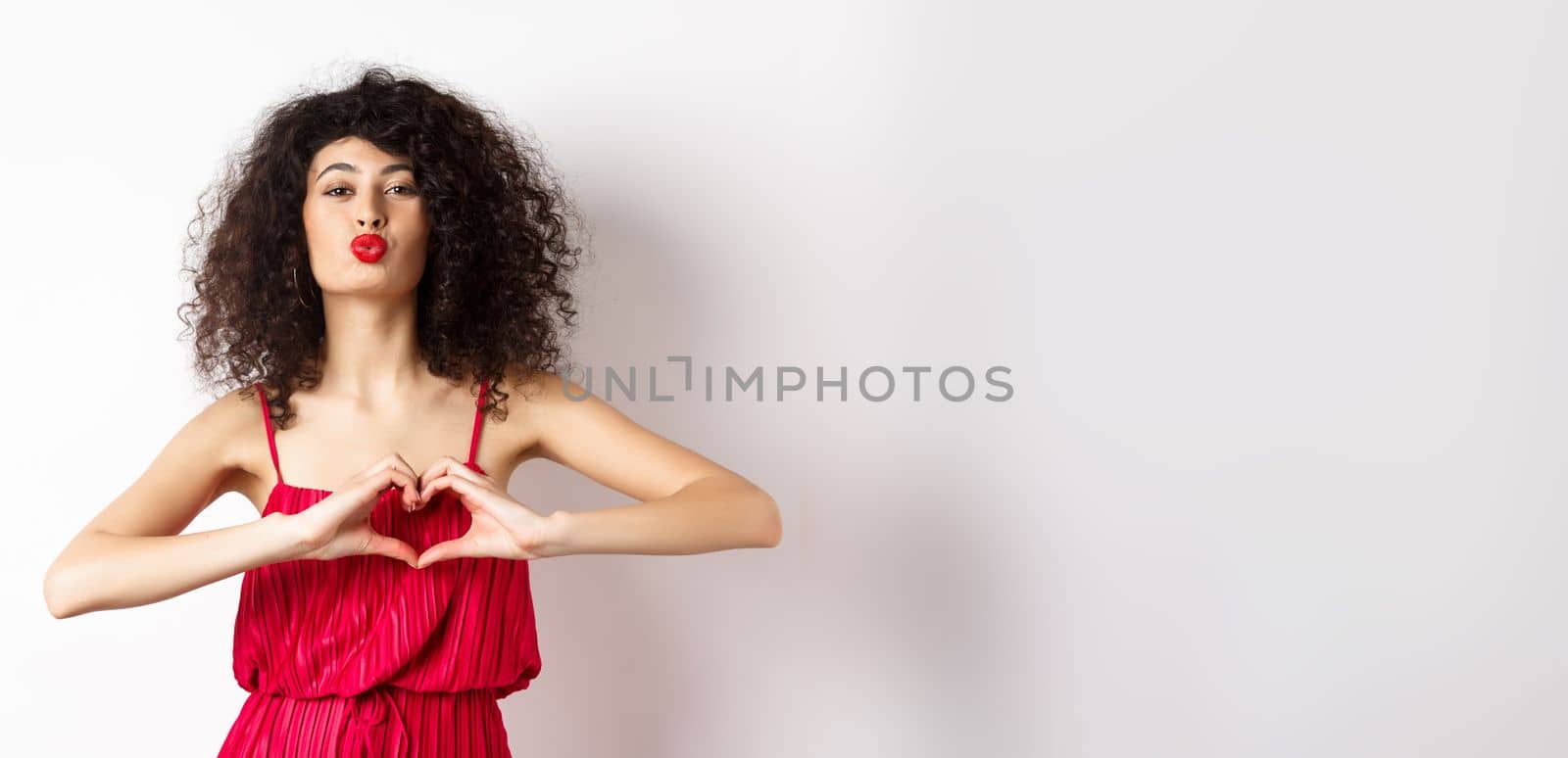Beautiful lady with curly hair, red dress, showing heart symbol and pucker lips for kiss, love you gesture, standing on white background by Benzoix