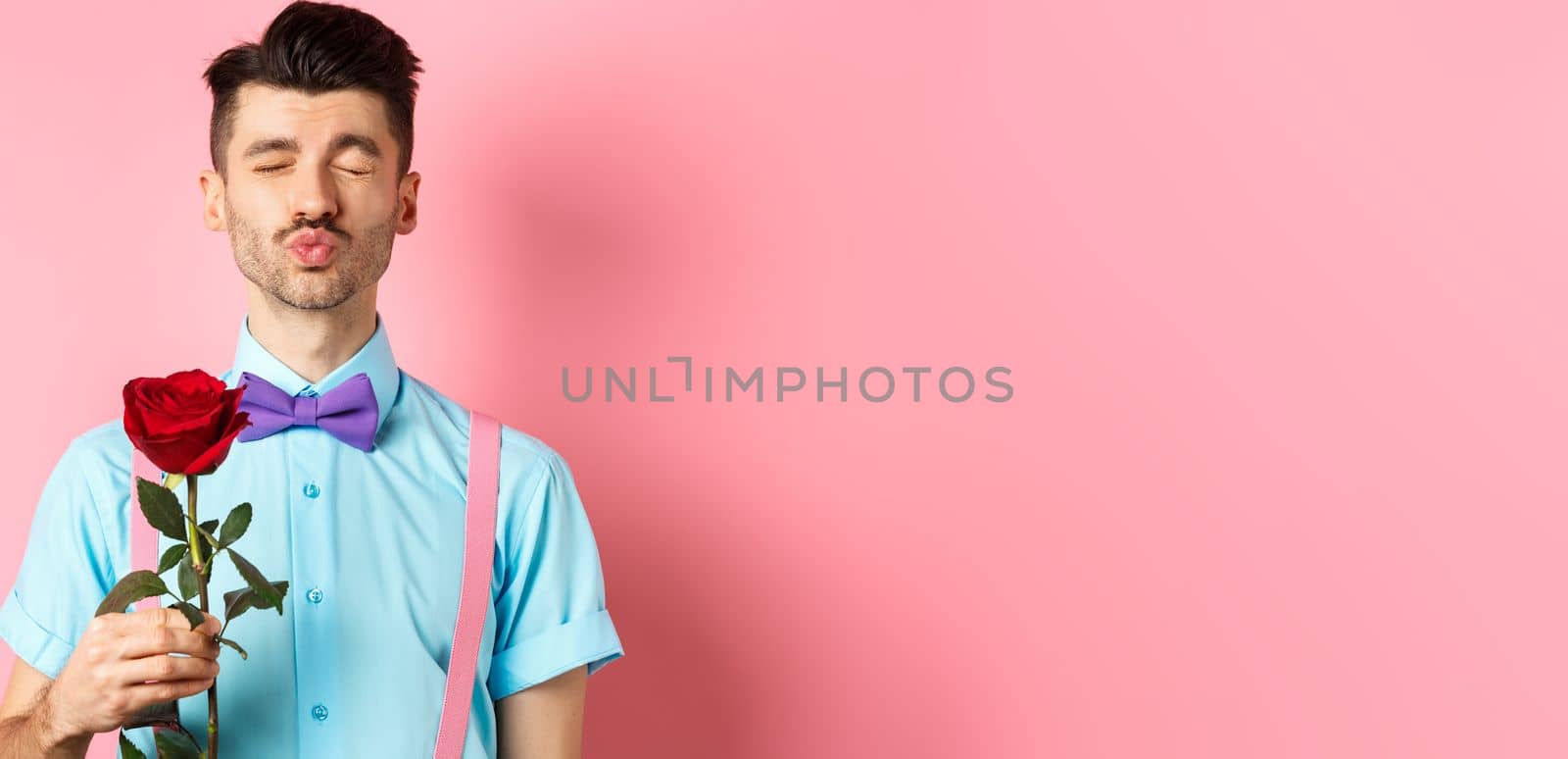 Cute and funny man waiting for kiss from lover on Valentines day, holding beautiful red rose for girlfriend, standing over pink background.