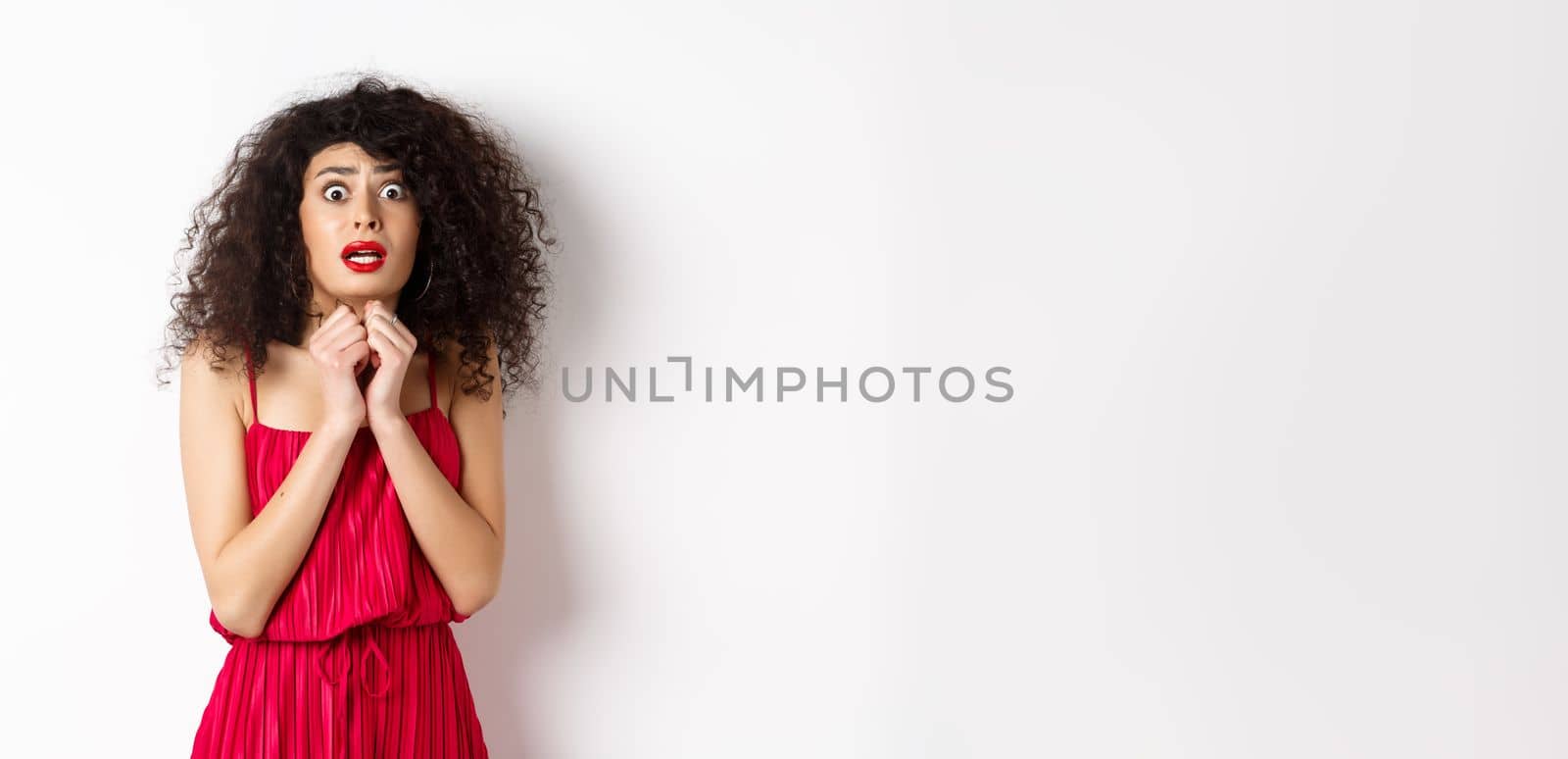 Scared caucasian woman trembling from fear, wearing red dress and staring anxious at camera, standing over white background.