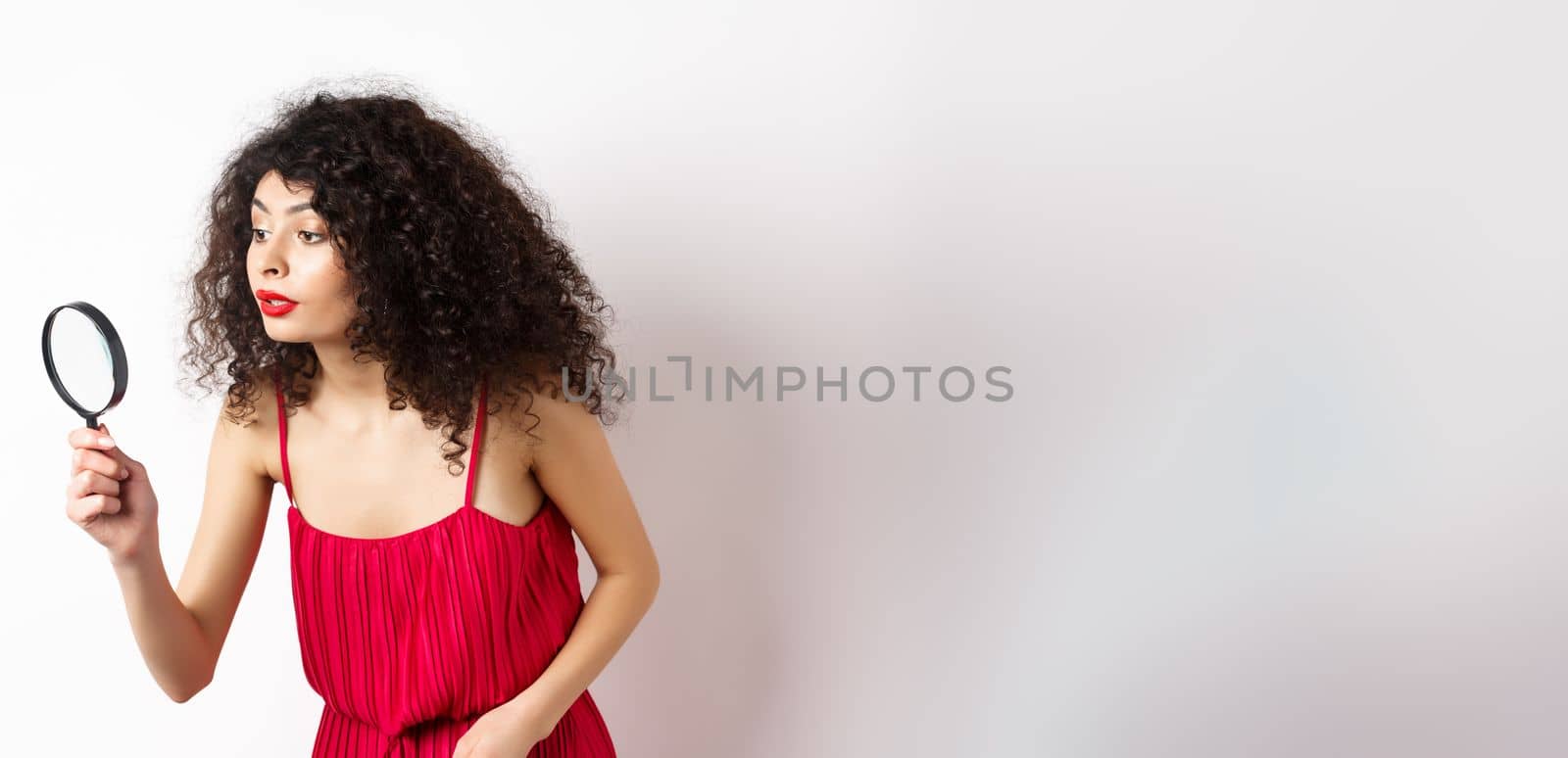 Beautiful woman in red dress searching for something, looking aside through magnifying glass, standing against white background by Benzoix