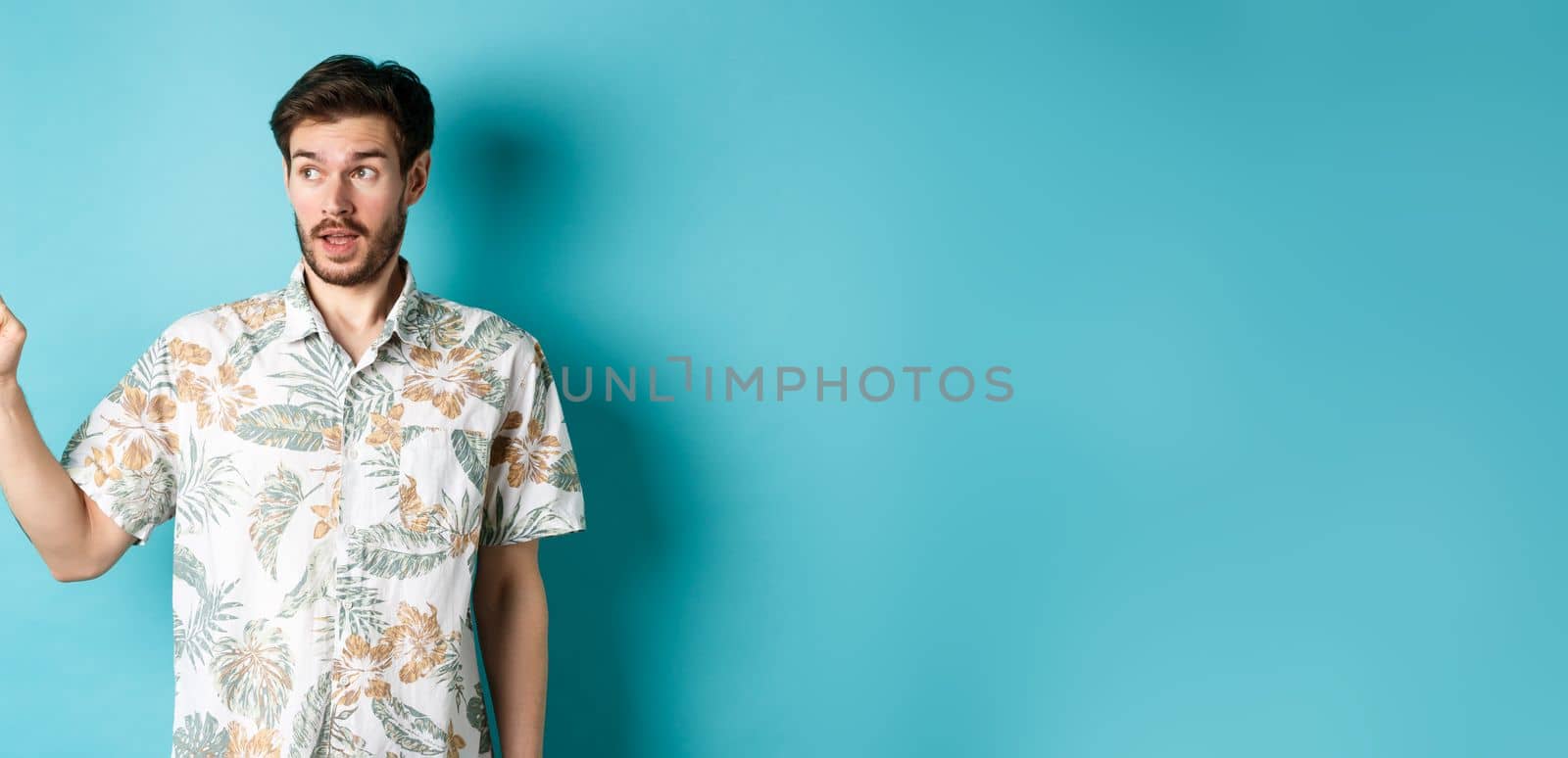 Intrigued man in hawaiian shirt, looking and pointing left at something interesting, blue background. Concept of tourism and vacation.