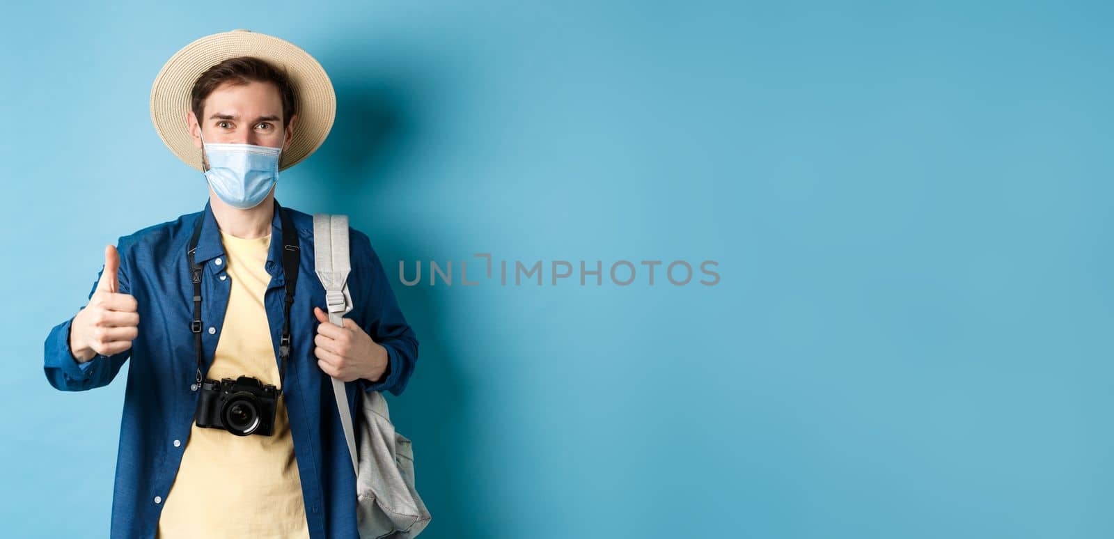 Covid-19 and summer holidays concept. Smiling guy travelling in medical mask and straw hat, backpacking and showing thumbs up, approve and praise travel agency, blue background.