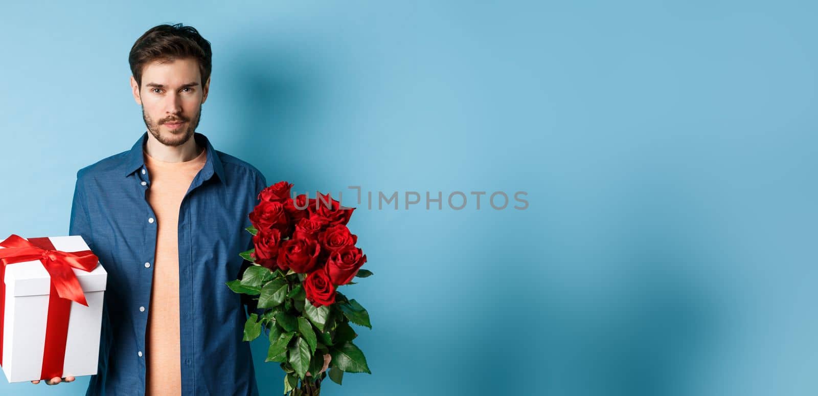 Love and Valentine day concept. Passionate man looking confident at camera, holding gift box and red roses for romantic date, standing over blue background by Benzoix