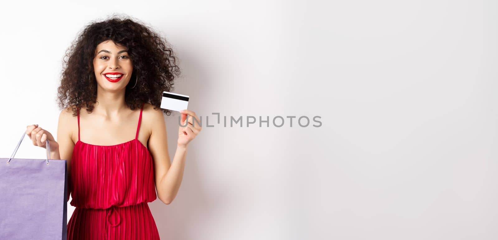 Beautiful female model with curly hair, red dress, showing shopping bag and plastic credit card, white background by Benzoix