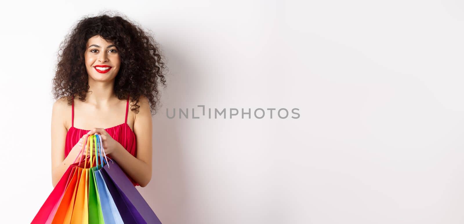 Image of stylish caucasian woman in red dress and makeup, holding shopping bags and smiling, standing over white background.