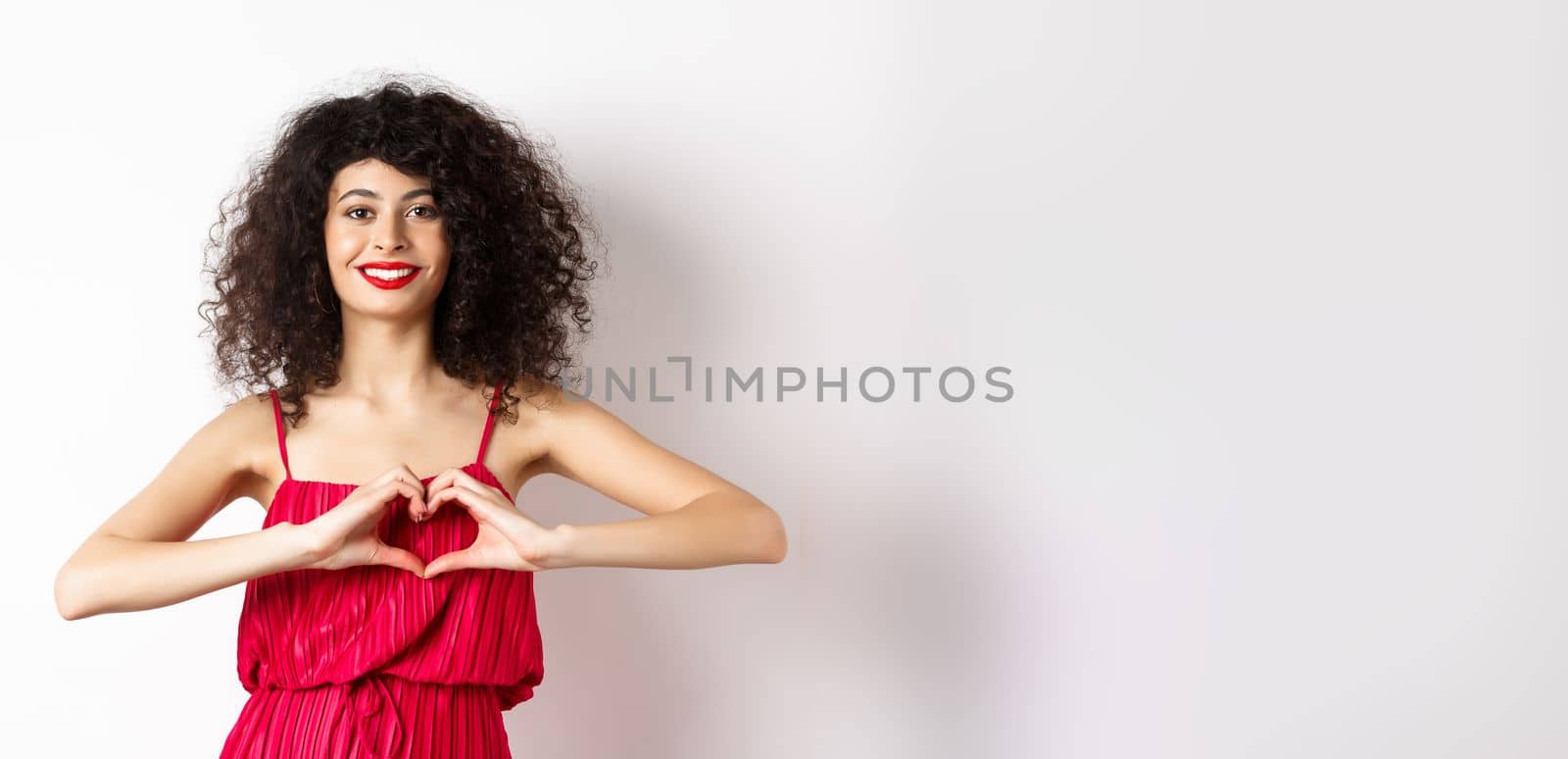 Charming young woman with red romantic fress, showing heart gesture and smiling, express love, standing over white background.