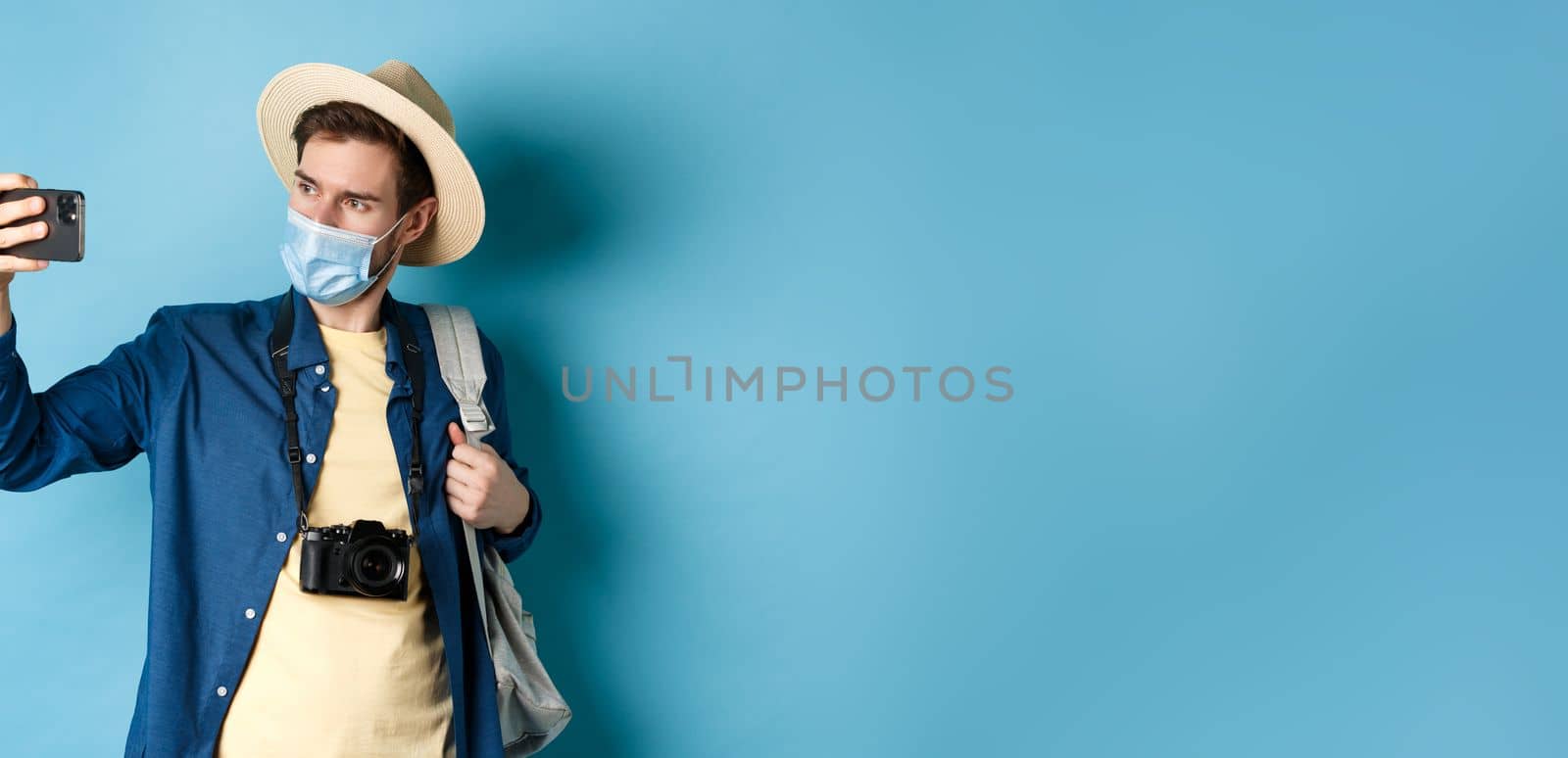 Covid-19, pandemic and travel concept. Male tourist in straw hat and medical mask recording video on smartphone during vacation, taking pics on summer holiday, blue background by Benzoix