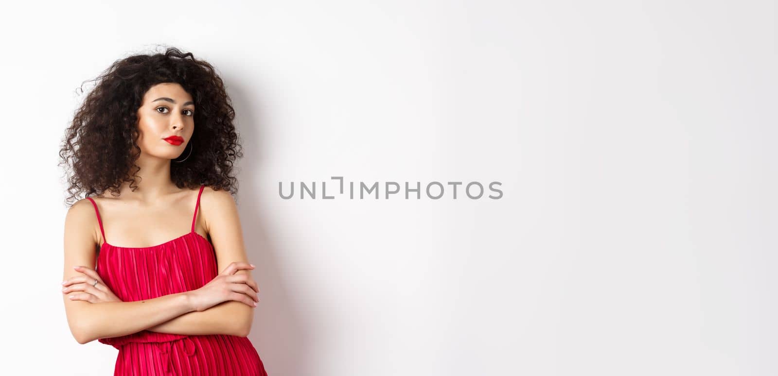 Sassy woman with curly hairstyle, wearing red dress and makeup, cross arms on chest, standing over white background.