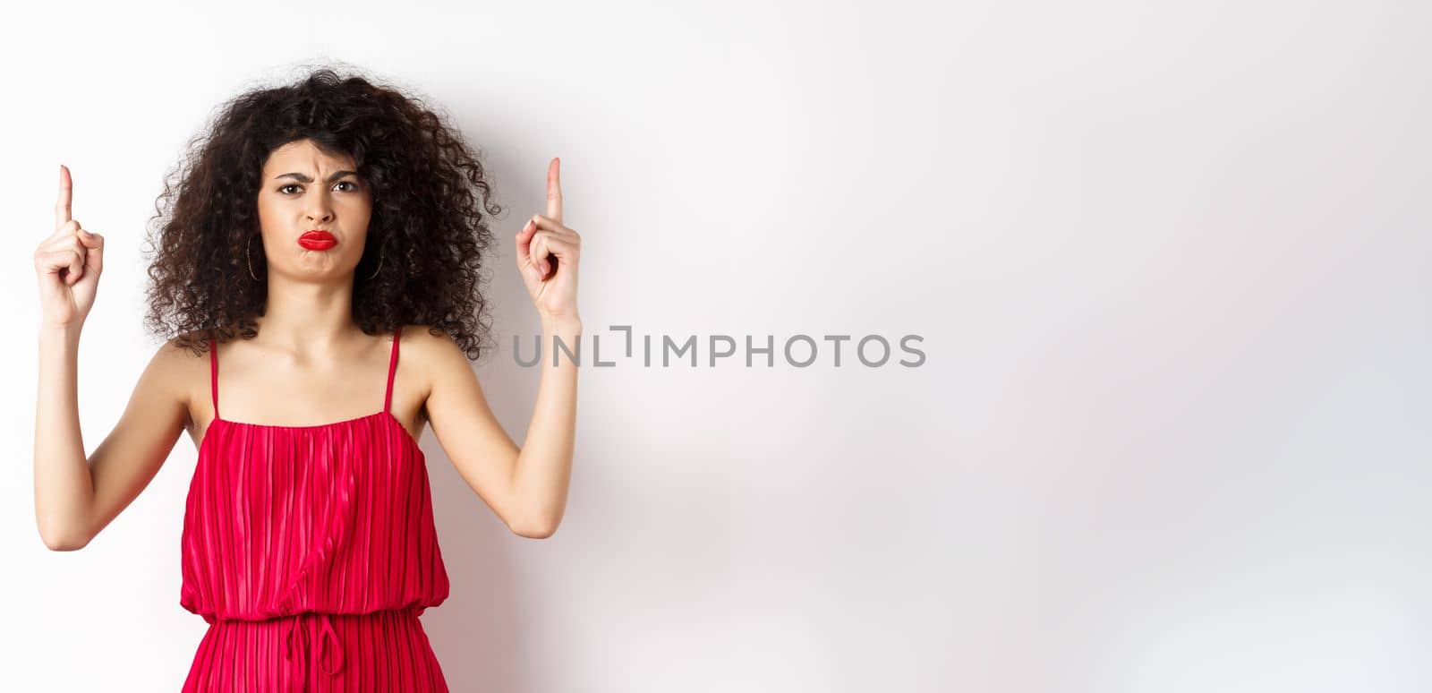 Disappointed frowning woman with makeup, wearing red dress on valentines, pointing fingers up and complaining, standing on white background by Benzoix