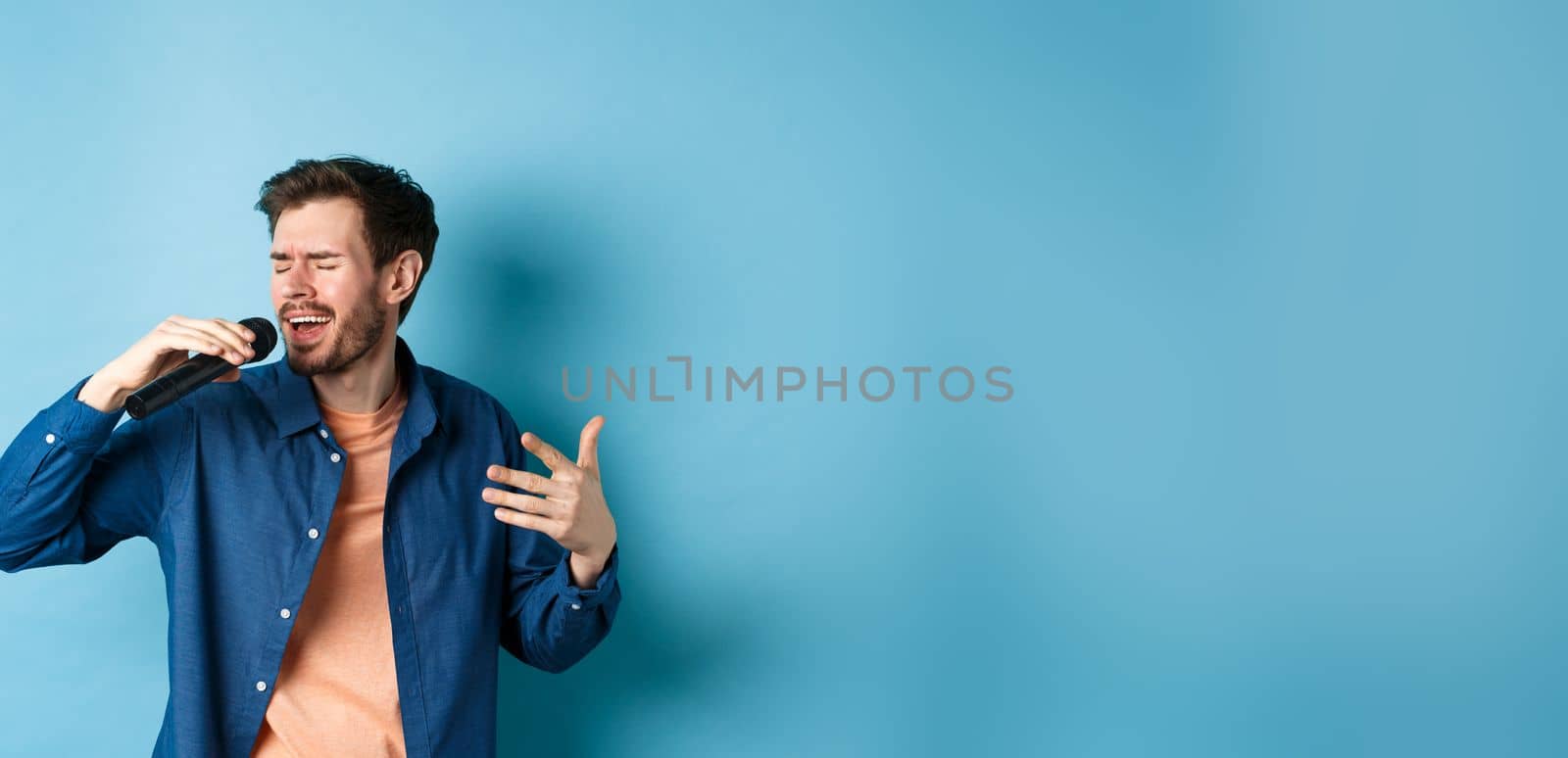 Romantic guy singing song in microphone and gesture, singer playing karaoke, standing on blue background.