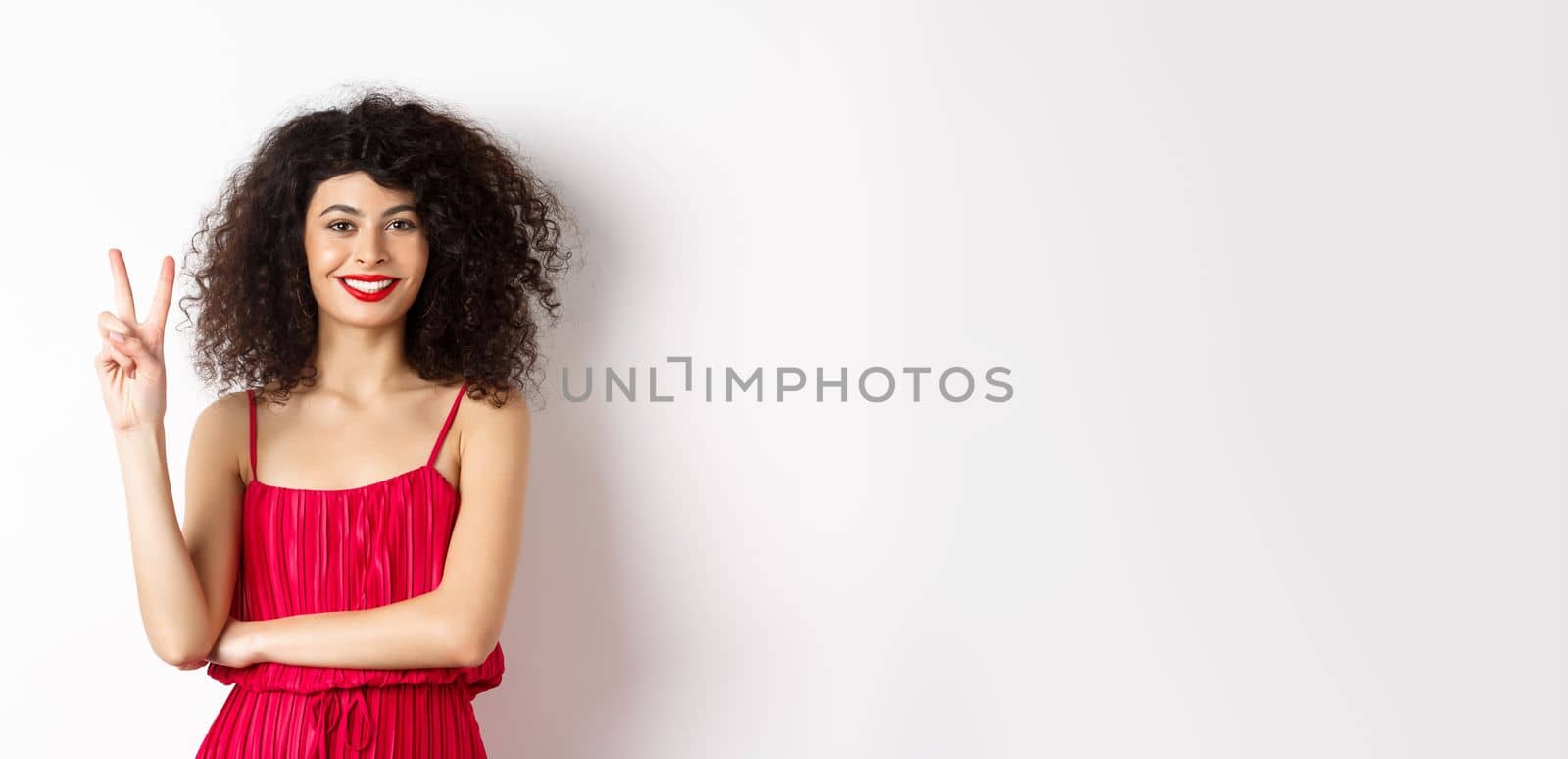 Attractive female model in red dress and makeup, showing number two and smiling, standing over white background by Benzoix