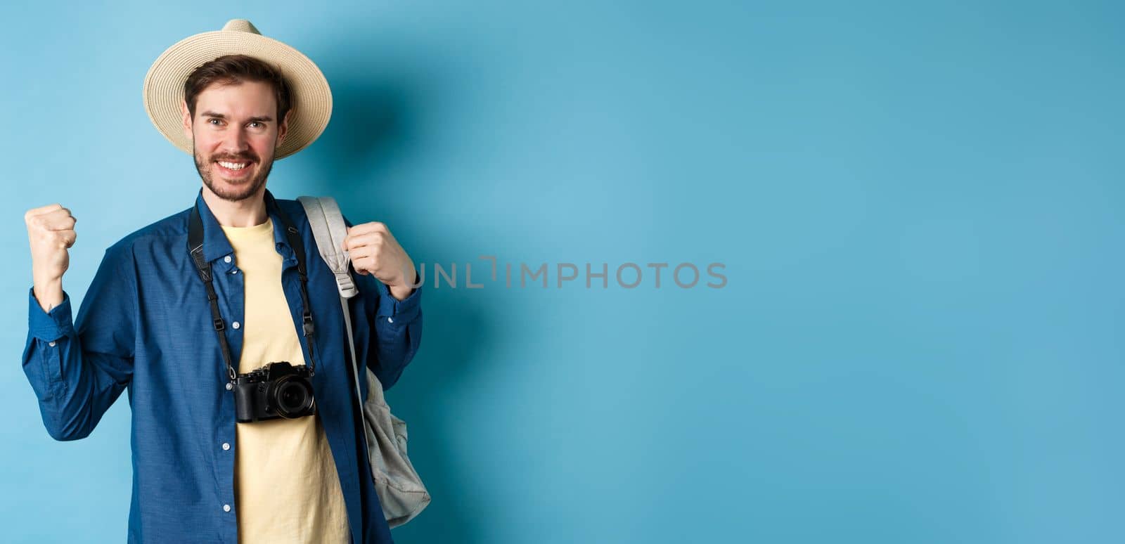 Happy tourist enjoying summer vacation, smiling and saying yes with fist pump, celebrating on holidays, feeling joy as finaly travel abroad, standing on blue background by Benzoix