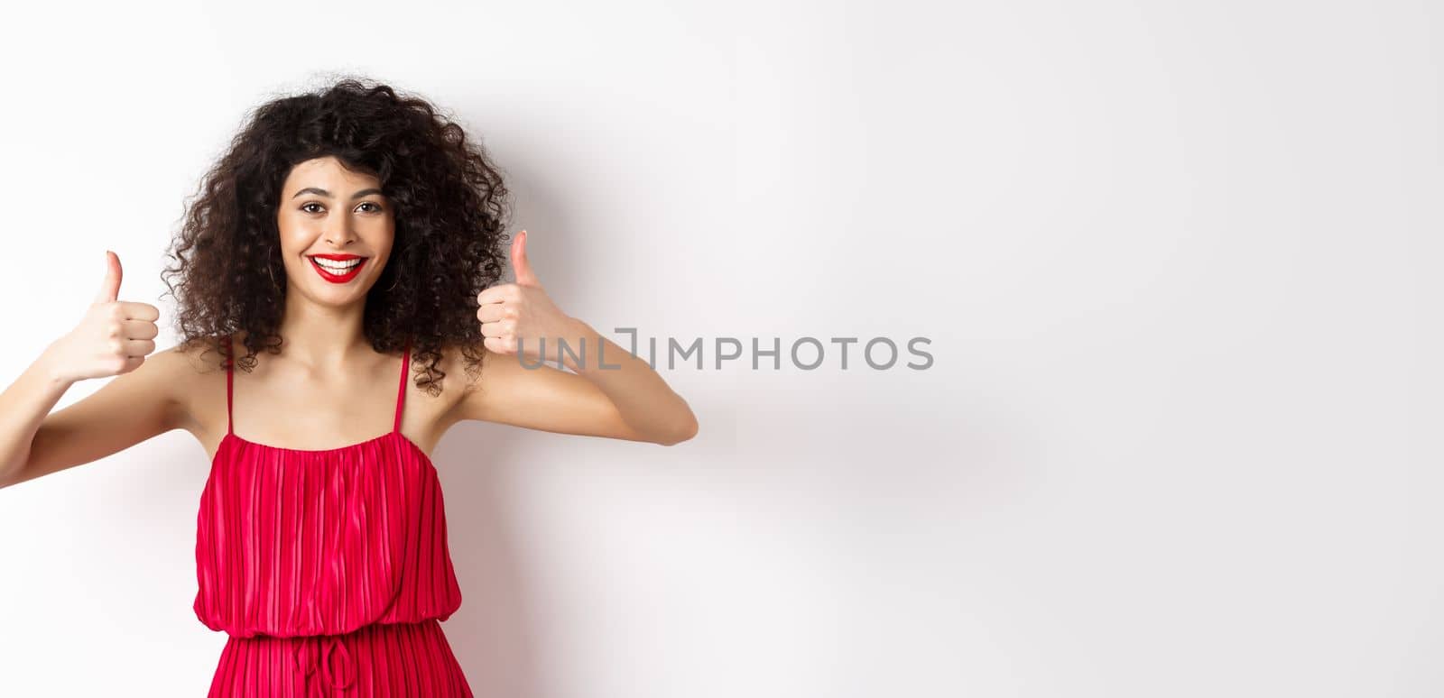 Attractive young woman recommending promo offer, showing thumb up and smiling, like product, standing in festive red dress on white background.