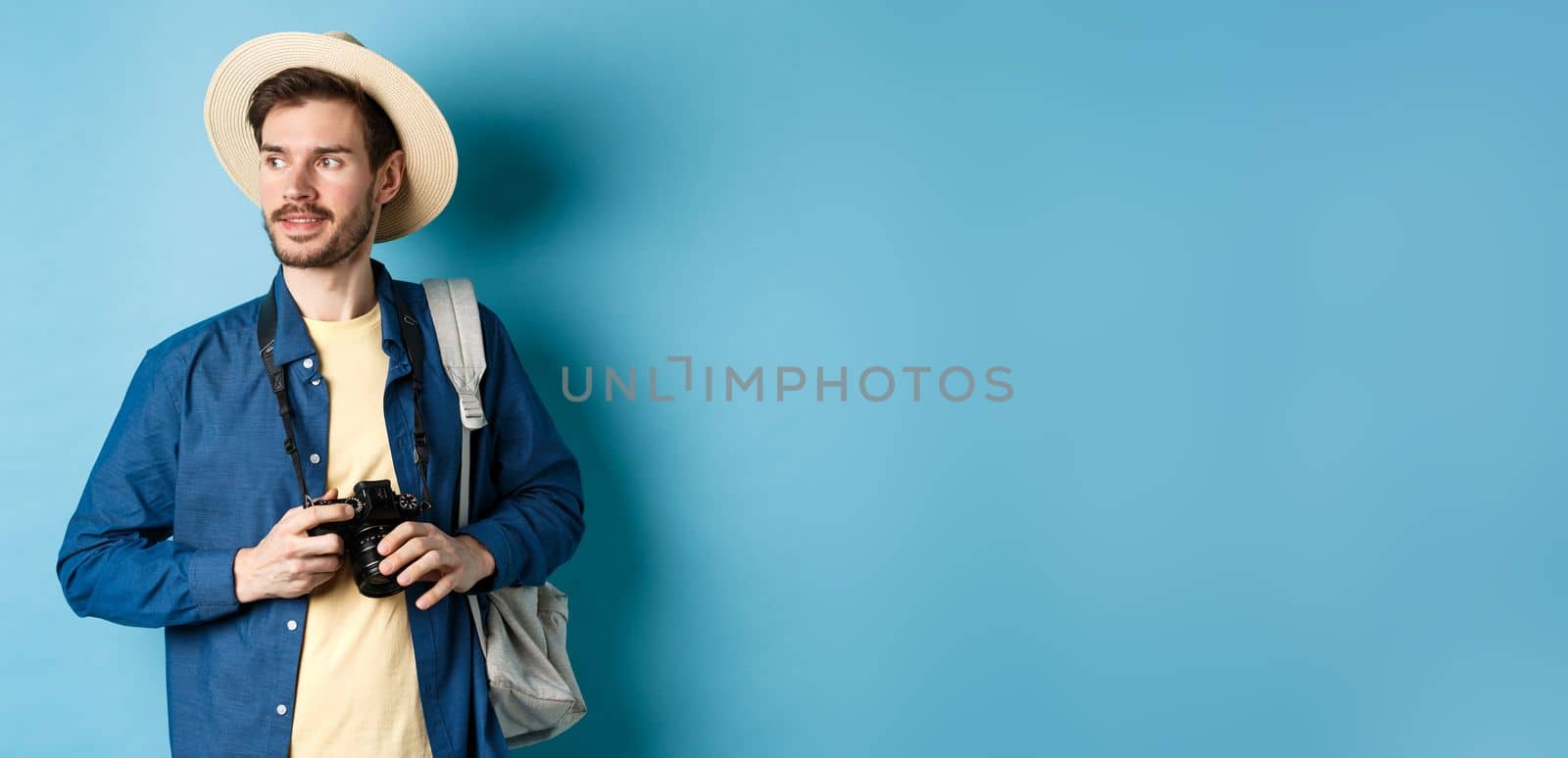 Handsome positive guy in summer hat, holding photo camera and looking aside, tourist taking pictures on vacation, standing with backpack on blue background by Benzoix