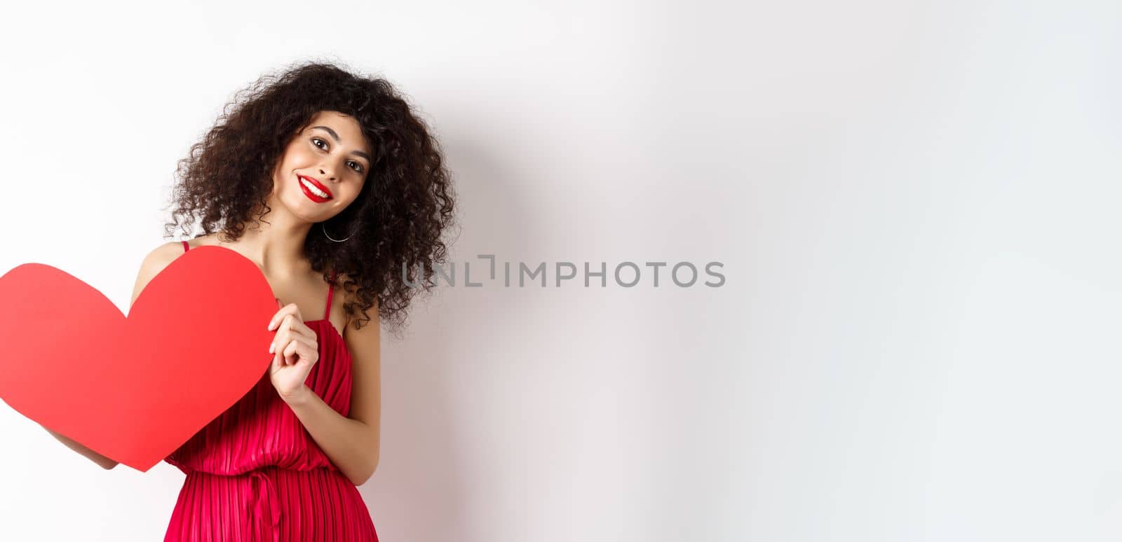 Romantic woman in dress showing big red heart, falling in love, smiling happy at camera, white background.