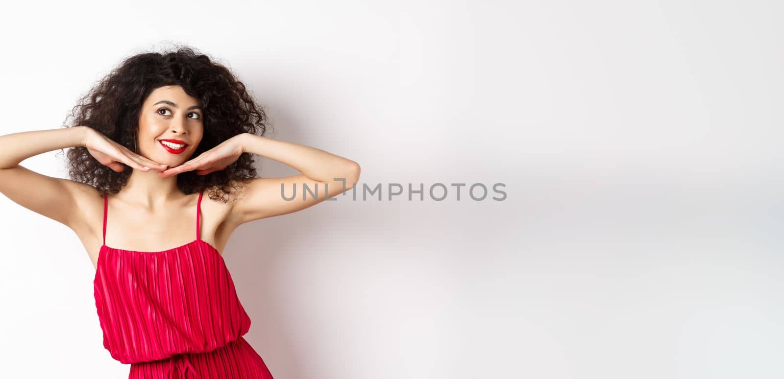 Beautiful lady with curly hair, wearing red dress, showing hear face with makeup and smiling, feeling carefree on white background.