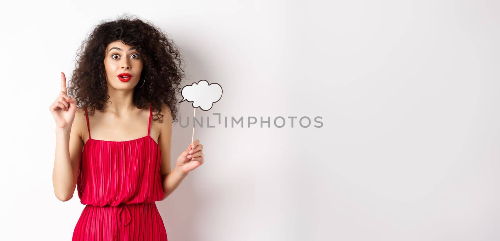 Cute caucasian lady in red dress, holding comment cloud and raising finger, pitching an idea and smiling, suggesting something, standing over white background by Benzoix