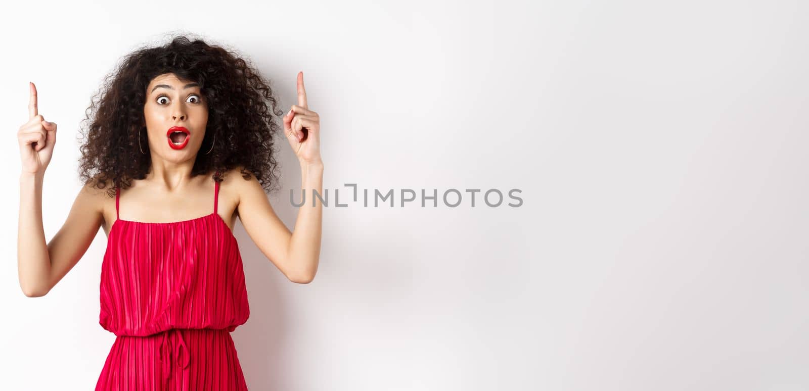 Impressed young woman with curly hair, wearing red dress, gasping and saying wow, pointing fingers up at logo, standing over white background by Benzoix