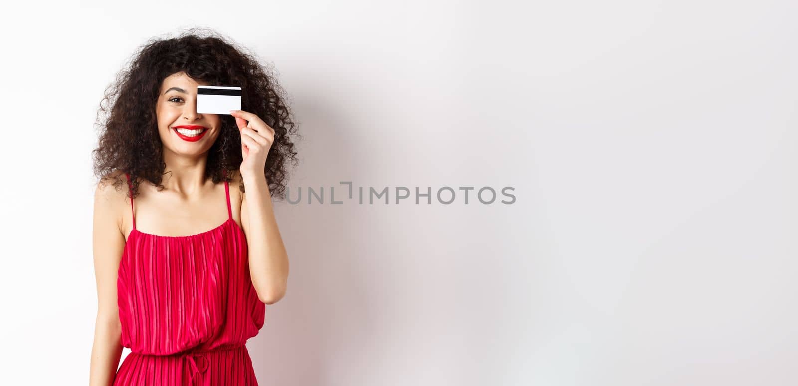 Image of elegant woman in red dress, showing plastic credit card and smiling at camera, standing over white background. Copy space