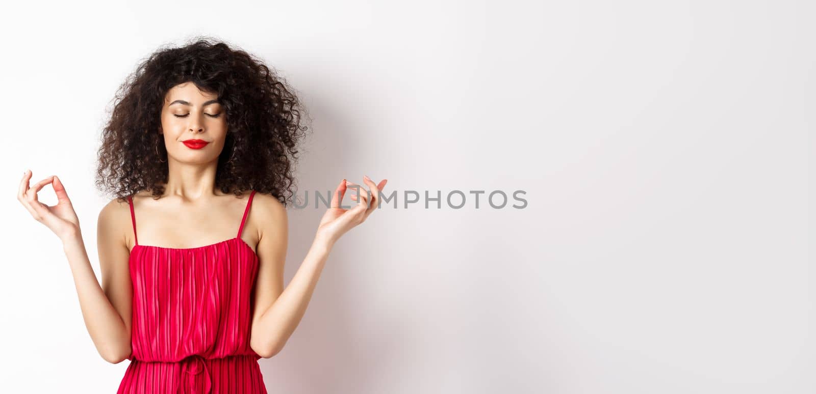 Calm and relaxed smiling woman in red dress, close eyes and meditating, practice yoga in zen pose, standing on white background.