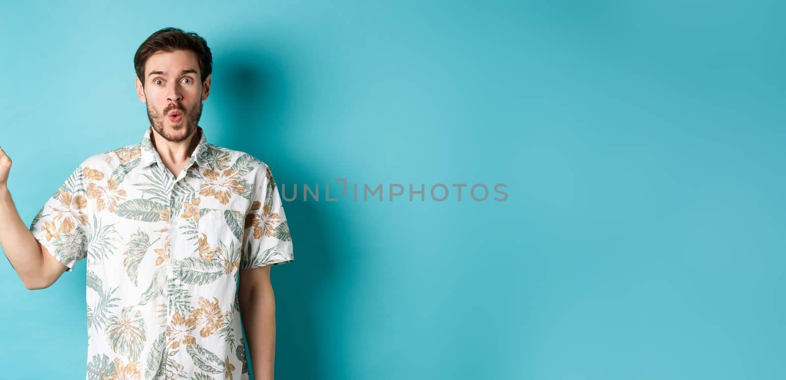 Surprised and amazed tourist in hawaiian shirt pointing finger left, showing promo and staring at camera, standing on blue background by Benzoix