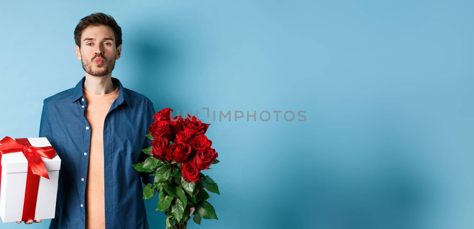 Love and Valentines day concept. Romantic boyfriend pucker lips for kiss, bring bouquet of red roses and gifts on date, standing over blue background by Benzoix