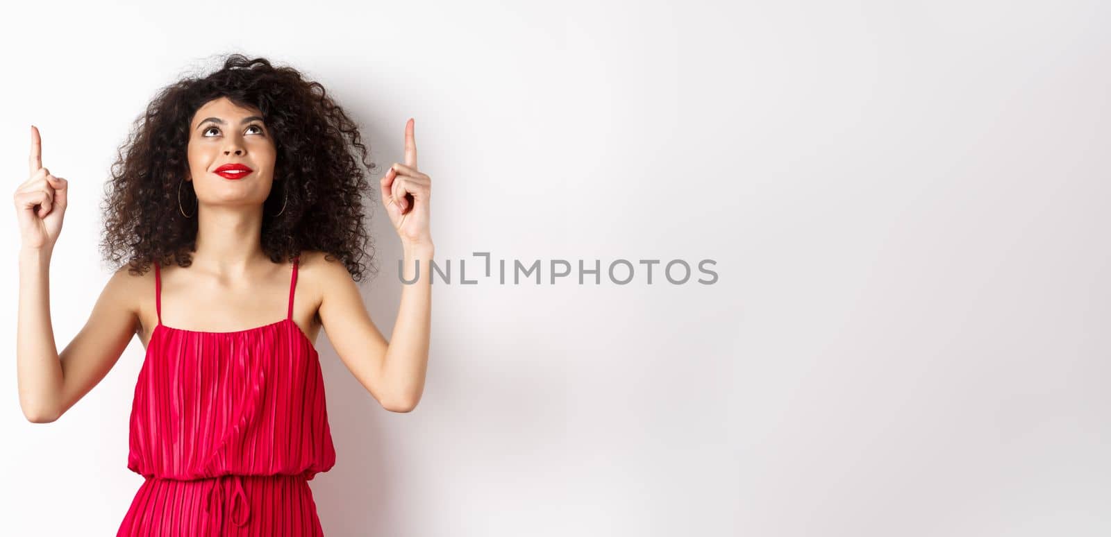 Dreamy young woman in trendy red dress, looking up and pointing at logo, smiling happy, standing over white background by Benzoix