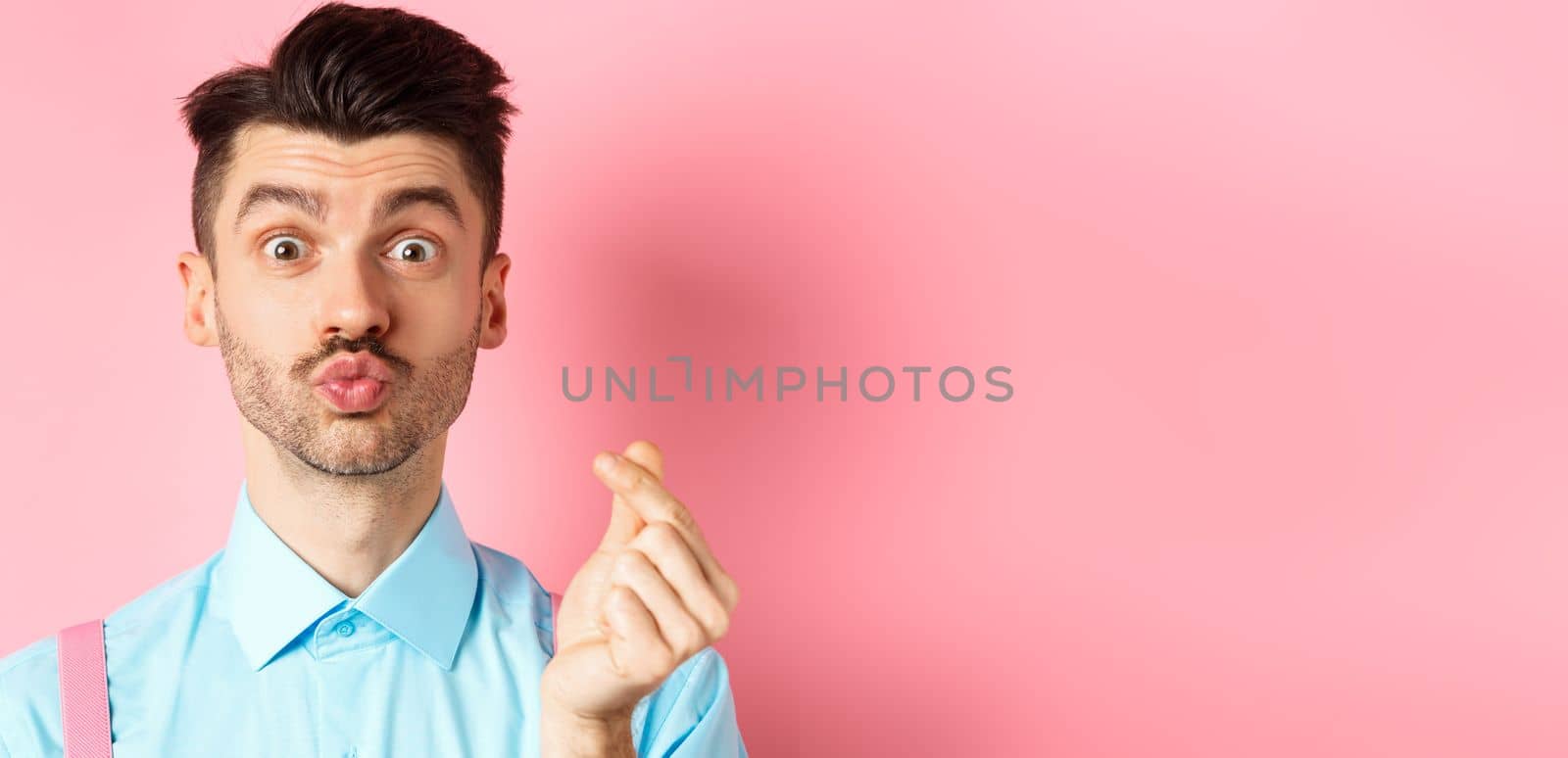 Valentines day concept. Silly guy waiting for kiss and showing finger heart gesture, express love and sympathy, pink background.