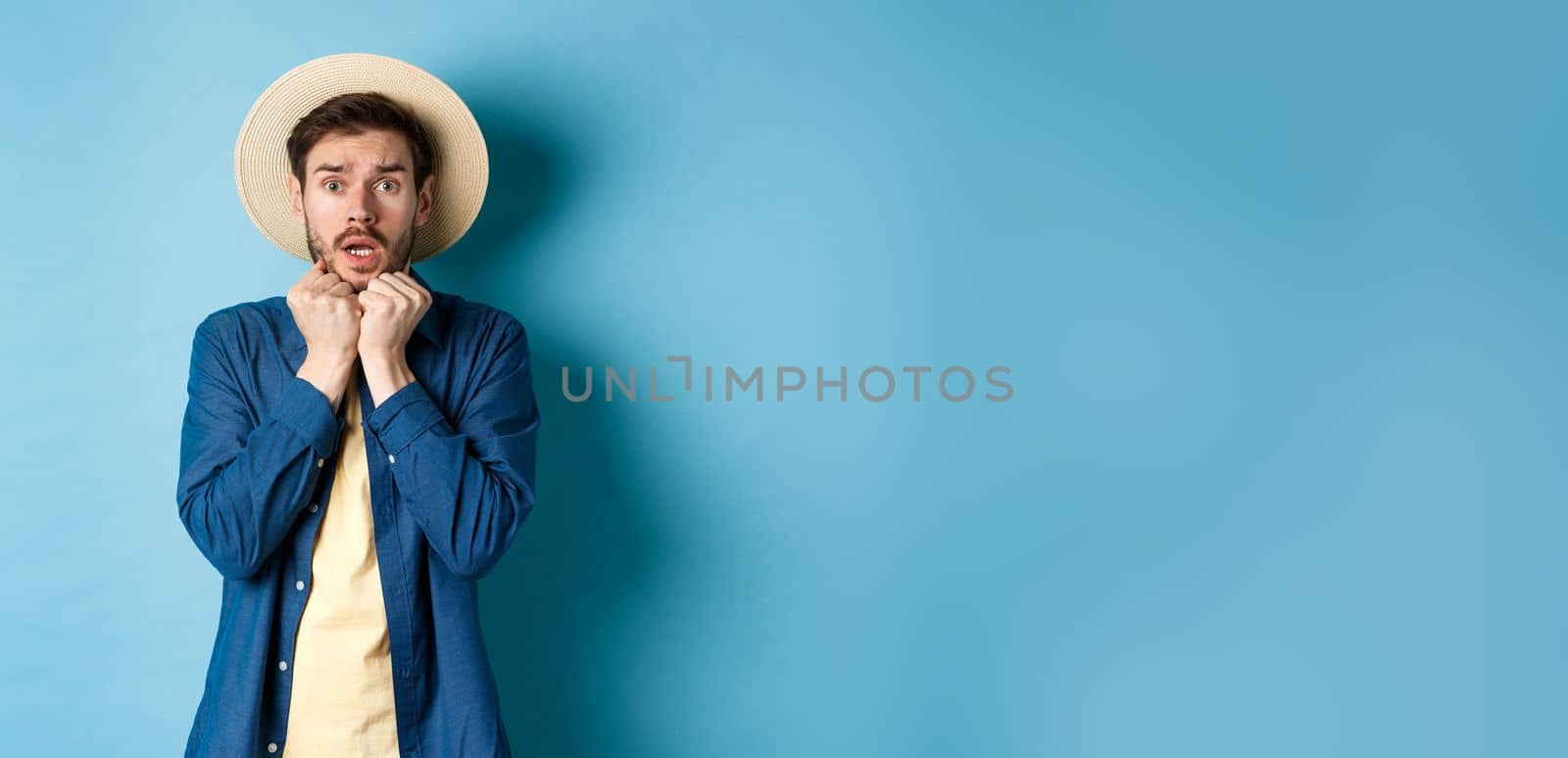 Scared young tourist trembling from fear, looking at something scary on summer vacation, wearing straw hat, standing on blue background.