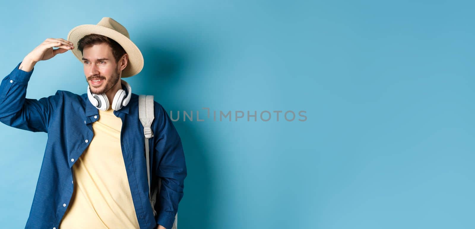 Handsome tourist in straw hat, covering face from sun and looking at distance aside, smiling pleased, travelling on summer vacation, standing over blue background by Benzoix