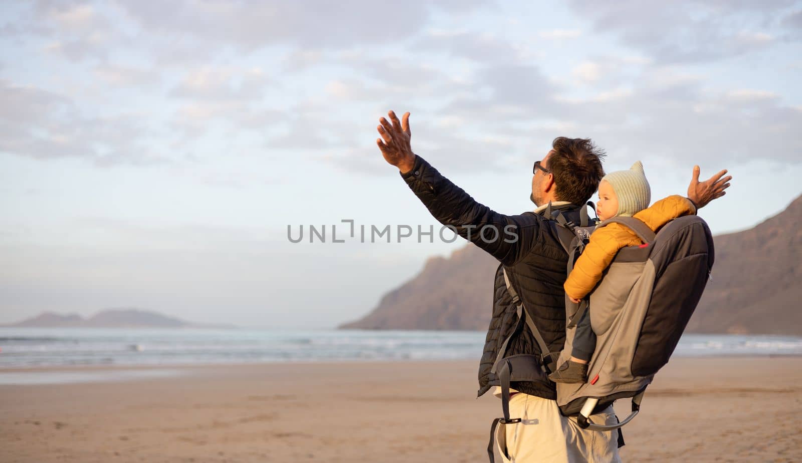 Father rising hands to the sky while enjoying pure nature carrying his infant baby boy son in backpack on windy sandy beach of Famara, Lanzarote island, Spain at sunset. Family travel concept