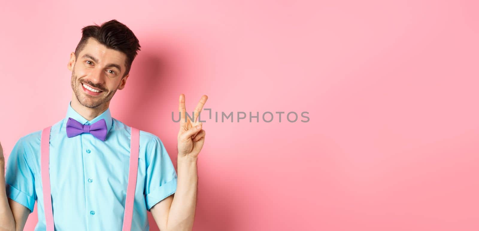 Romantic guy in bow-tie and suspenders showing peace signs and smiling at camera, wearing clothes for date, standing on pink background.