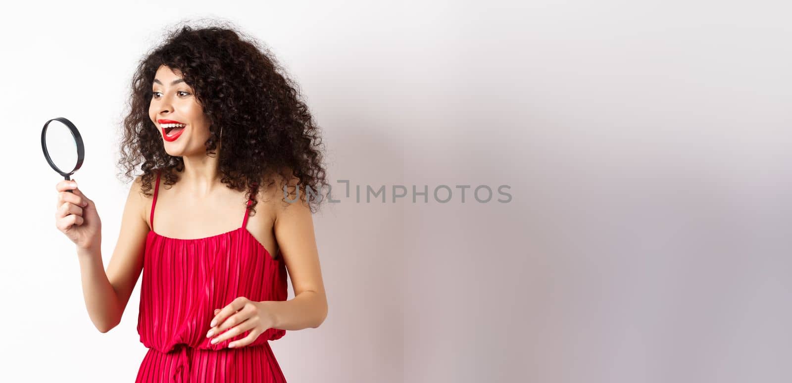 Excited woman in red dress look through magnifying glass and smiling, found interesting promo, standing on white background by Benzoix