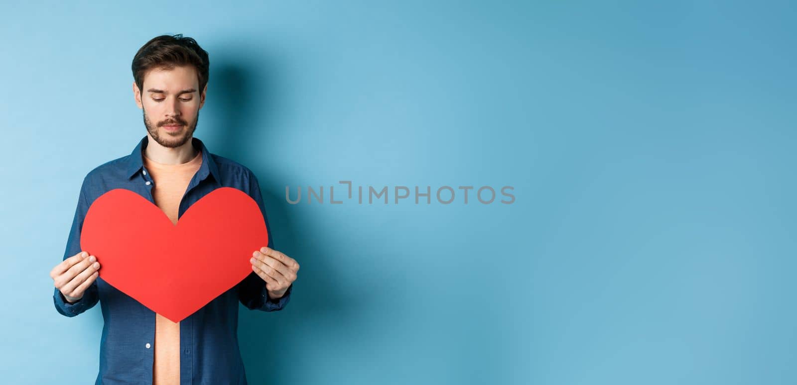 Lonely guy looking sad at valentines red heart with sad face, standing over blue background.