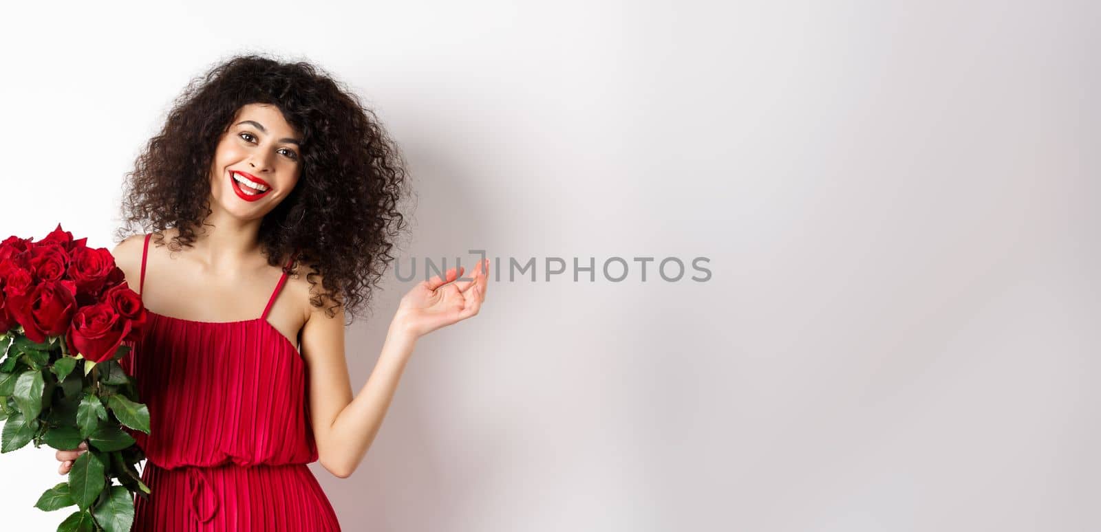 Happy woman celebrating, wearing stylish dress and holding flowers, smiling at camera, white background.