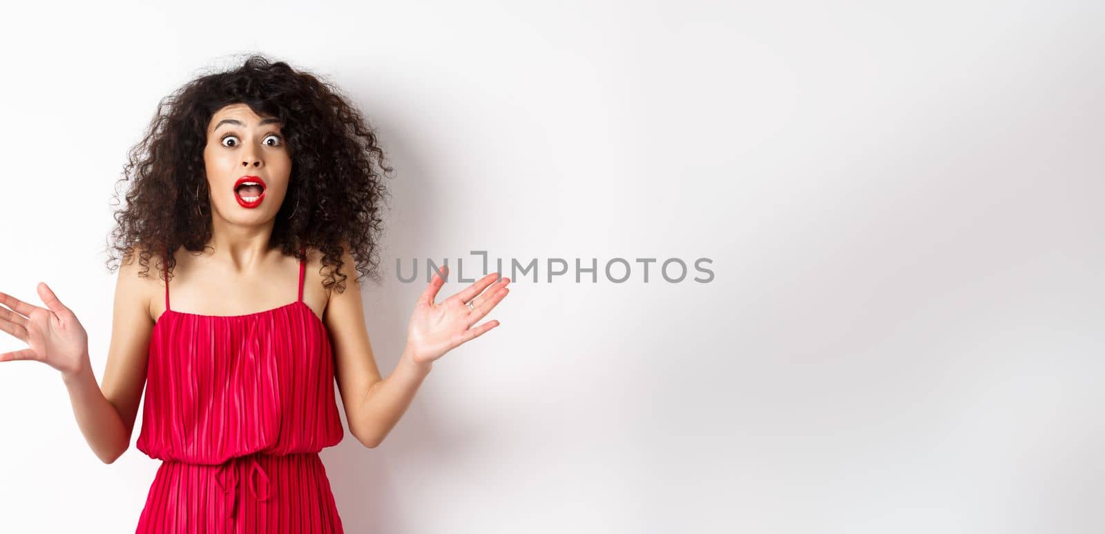 Excited woman with curly hair, talking with amazed face, explain big news, shaking hands and look astonished at camera, white background.