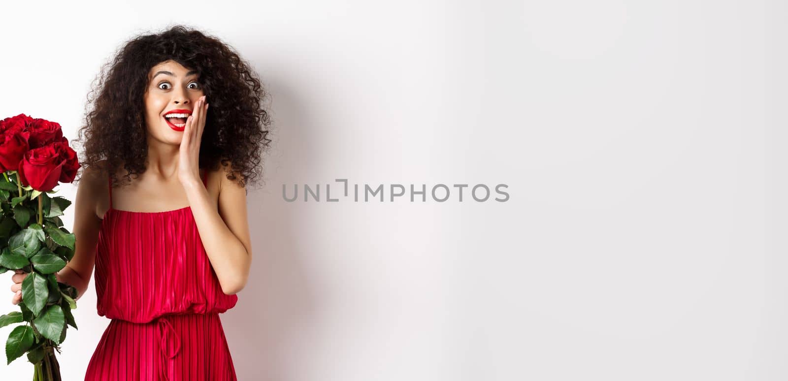 Excited curly woman in red dress, receive bouquet of roses and look surprise, rejoicing from romantic gift, standing on white background by Benzoix