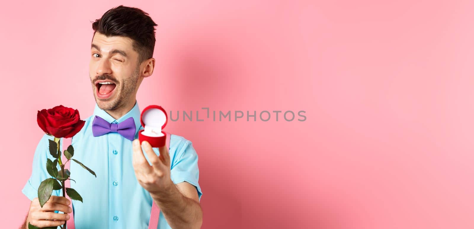 Valentines day. Funny guy making proposal, winking and saying marry me, showing engagement ring with red rose, standing over pink background.