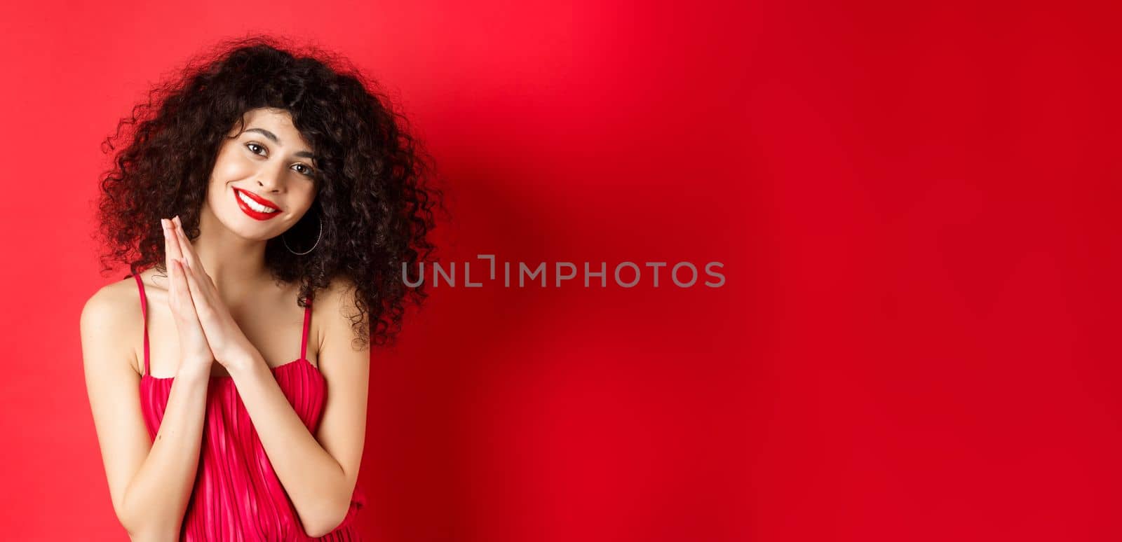 Tender woman with curly hair, wearing red dress, looking at something lovely, thanking you, standing over studio background. Copy space